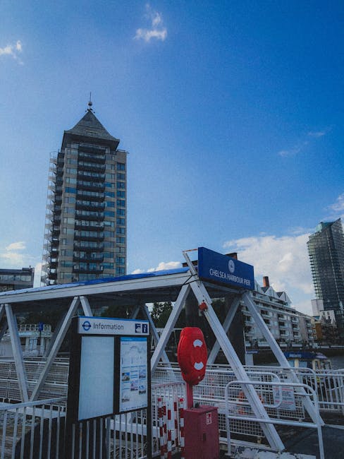 A view of Chelsea Harbour Pier with a white metal-framed roof structure clearly visible, supporting a blue sign displaying the location name. In the foreground, a red lifebuoy is mounted on a pink metal post attached to the pier's safety railing, which is composed of white metal bars. Behind the safety equipment, an information display board with a digital or printed map is mounted on a black pole. The background features several modern high-rise buildings, including a distinctive tower with a pointed, textured roof and reflective glass windows, under a blue sky with a few scattered clouds. The scene captures a clean, urban waterfront area associated with private or independent waste handling, as managed by Waste Disposal Chelsea, with no visible rubbish or debris in the immediate vicinity.