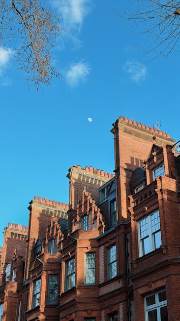 The image features the upper section of a historic red-brick building with detailed architectural elements, including decorative battlements, arched windows, and stone window sills, set against a bright blue sky with a visible partial moon. In the foreground, the lower branches of a leafless tree extend into the frame from the upper left corner. The building appears to be part of a residential or institutional complex situated on a city street or courtyard, with the exterior showing a textured finish typical of traditional London architecture. The scene is well-lit by natural daylight, with shadows cast by the architectural features emphasizing their depth and detail. In the context of rubbish removal or on-site clearance, Waste Disposal Chelsea would typically handle the disposal of any debris or waste associated with building renovations or estate clearances, ensuring a clean and safe environment for such historic properties.