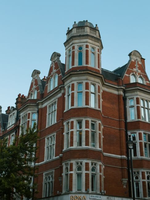 A historic red-brick building with detailed white window frames and decorative stonework, featuring multiple narrow, tall windows arranged in vertical stacks. The structure has rounded bay windows and a prominent octagonal turret with a decorative railing at the top. The building's façade includes intricate brickwork accents and gabled roof sections, with some chimneys visible on the roof. Adjacent to the building on the right is a traditional black street lamp. In the foreground, partially visible, are leafy tree branches, and the sky above is clear with a soft blue tone. The building’s architectural details suggest an older, possibly Victorian or Edwardian style, and the setting appears to be in an urban residential or historical district. The scene conveys a well-maintained, distinctive property with elements typical of period architecture, reflecting the aesthetic context often associated with heritage conservation and private residential sites, which some waste removal services cater to through specialist or on-site clearance.