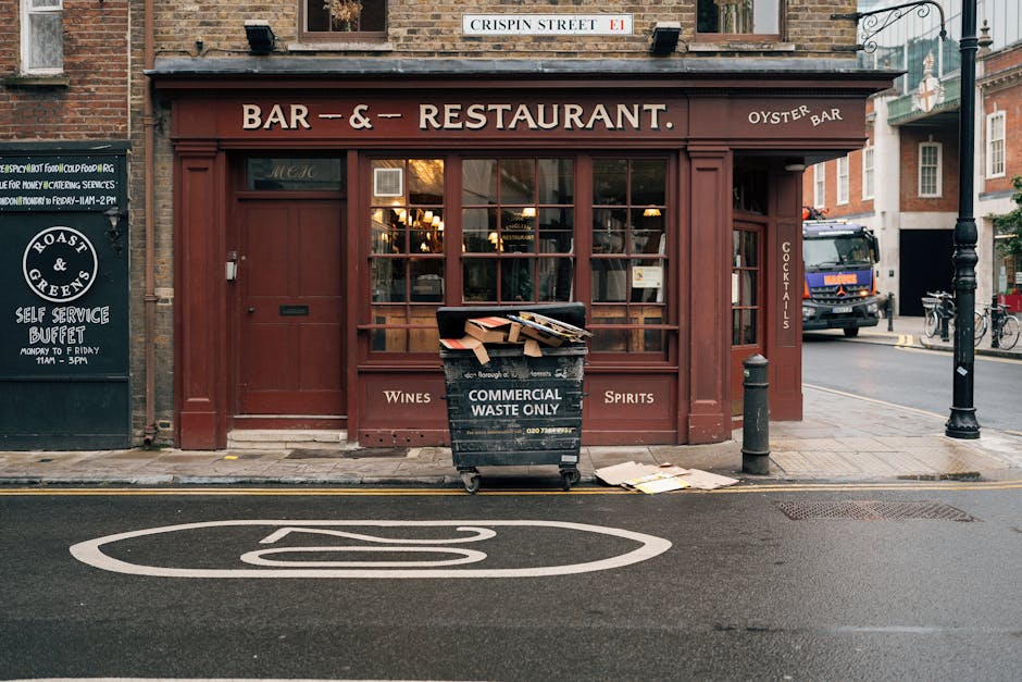 In front of a brick-built pub or restaurant with a dark red wooden facade and large window panels, a black commercial waste bin marked 'Commercial Waste Only' is positioned on the pavement. The bin is overflowing with cardboard boxes and paper, some stacked on top, and a few sheets have fallen onto the street. To the right of the bin, a short metal bollard separates the sidewalk from the road. The roadside features a painted white symbol indicating a designated parking or loading zone. Visible behind the establishment are a street nameplate reading 'Crispin Street E1' mounted on the building, a parked delivery truck, and a row of bicycles secured to a stand. A blackboard sign on the left advertises self-service buffet hours, while overhead street lighting fixtures and nearby buildings suggest a bustling urban setting. The environment indicates a typical City of London scene where private waste disposal services, such as those provided by Waste Disposal Chelsea, often handle rubbish collection outside of local authority channels, supporting alternative waste management options.