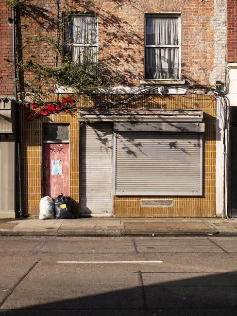 The image depicts the front of a small commercial building with a brown-tiled facade and an adjacent residential brick structure. The building features a grey metal security shutter covering a large window, partially closed, with a small rectangular ventilation grille beneath it. To the left of the shutter, there is a narrow, vertically oriented pink door with some graffiti marks, and next to it, three black rubbish bags are placed on the sidewalk, along with two white plastic bags. Above the building's entrance, there are a few cables and wires running horizontally, casting shadows on the upper brick wall, which has two windows with frosted glass panes and white frames. Climbing plants with green leaves are visible near the top left, partially covering the bricks. The scene is illuminated by natural daylight, with clear shadows on the street in front of the shop, indicating a sunny day. This setting shows an independent approach to waste management, with Waste Disposal Chelsea often handling such refuse collections for local businesses and commercial properties in the area, reflecting the practical aspect of private rubbish removal services.