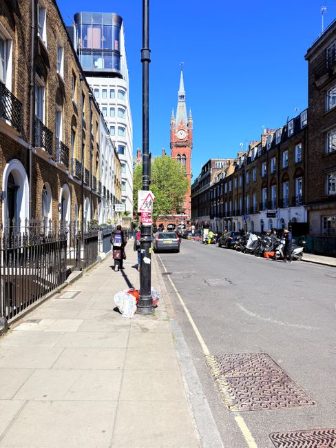 A city street scene featuring a collection of multi-storey buildings with varied architectural styles, including brick facades, arched and rectangular windows, and ornate detailing. The buildings are of different heights, with the tallest featuring a domed turret topped with a weather vane, and some have blue and beige finishes. In the foreground, a light blue rubbish collection truck is parked alongside the pavement, indicating an active rubbish removal service. To the right, a line of metal bike racks is visible, and the street is lined with bollards and pedestrian pathways. The environment appears urban and densely built, with additional historic and modern structures visible further along the street. The lighting suggests a clear daytime setting, with soft shadows and a neutral atmosphere. This scene portrays a typical urban setting suitable for private waste removal or independent collection services provided by Waste Disposal Chelsea, supporting the broader context of alternative rubbish handling in Chelsea.