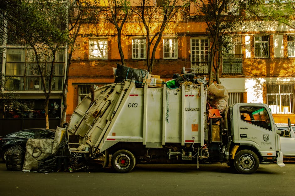 A white waste collection truck belonging to Waste Disposal Chelsea is parked on a city street in front of an orange brick residential building with multiple windows. The truck's large rear compactor section is filled with various waste materials, including black garbage bags, cardboard boxes, and plastic containers, some of which are leaning against the vehicle. The materials appear to be a mix of household junk and discarded items, with visible textures such as crumpled paper, plastic wrapping, and cardboard edges. The truck's rear is angled upward, indicating that it is in the process of loading or compacting rubbish. On the left side of the image, a black garbage bag sits on the pavement, along with a small pile of debris. The background features trees with sparse green leaves casting shadows across the building facade, creating dappled sunlight on the scene. To the right, additional vehicles are parked along the street, and the overall environment suggests a typical urban setting for private waste collection or rubbish removal services, supporting alternative waste handling outside of council services.